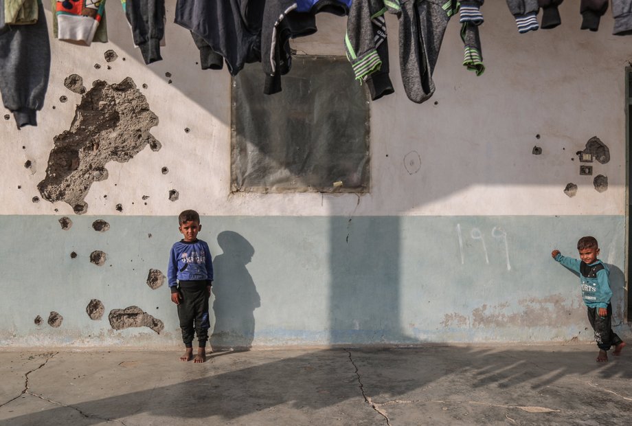 Syrische Kinder spielen vor einem Haus, dessen Wand durch Einschusslöcher beschädigt ist.