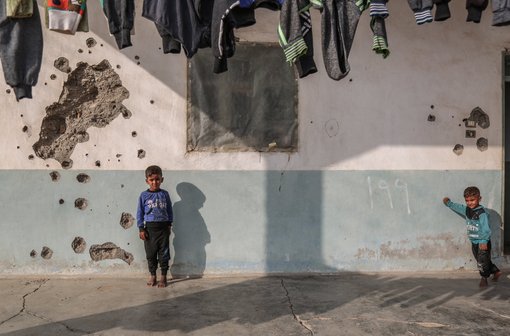 Syrische Kinder spielen vor einem Haus, dessen Wand durch Einschusslöcher beschädigt ist.